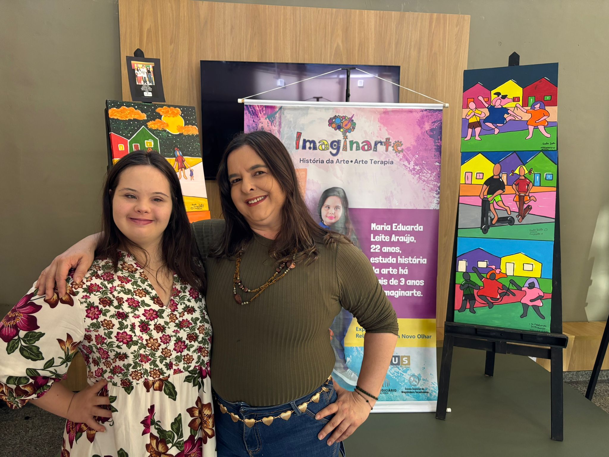 Na imagem, a jovem artista Maria Eduarda Leite (Duda) posa abraçada com sua professora de artes, Edineia. As duas estão sorrindo e com a mão na cintura. Duda, mulher branca com cabelos lisos e longos, veste um vestido florido de manga longa. Edineia usa blusa verde escura, colares e calça jeans azul escuro. Ao fundo, estão expostas as obras de arte coloridas produzidas por Duda, além de um banner com o texto: “Imaginarte — História da Arte • Arte Terapia. Maria Eduarda Leite Araújo, 22 anos, estuda história da arte há mais de 3 anos na Imaginarte”.