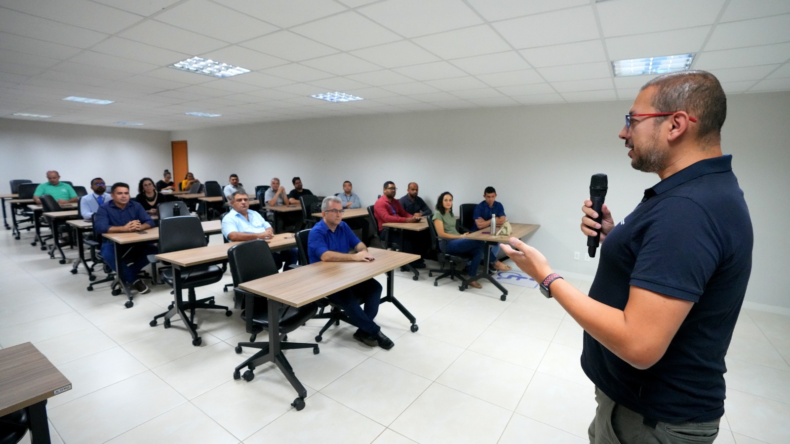 Em uma sala de aula, o professor está de perfil, com barba, óculos e camisa polo, segura um microfone enquanto fala para os alunos. Ele está em pé, voltado para os participantes, que estão sentados em carteiras organizadas em fileiras. A maioria dos participantes é composta por homens, e alguns têm notebooks ou garrafas sobre a mesa. 